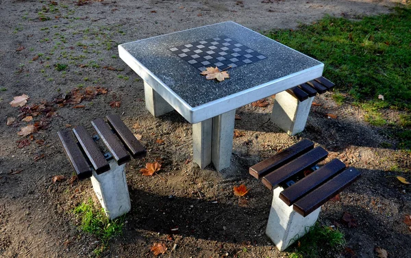 stock image Park furniture chess table and chairs for four persons made of light metal and wooden beams with backreston bright glade area gravel lawn green beige gray evening sun spring