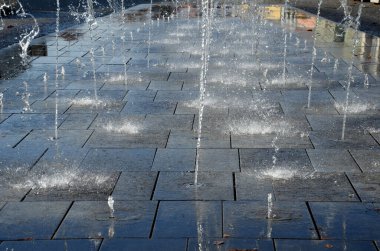 Kaldırımda kaldırıma yığılmış kare granit kaldırım taşları, sırayla püskürtülmüş su püskürtüleri. Su özellikleri doğrudan park sırasındaki kaldırımlı meydandan ortaya çıkıyor. Islak kiremitler tahta kaldırım, bebeğim, kep