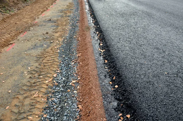 stock image A newly built road, on the layer of which you can see successive layers of screed and asphalt coatings and penetration spraying. the edge is gradually shaped by earth at ditch, uncomplete, fall down 