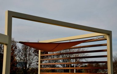 Sarı tuval gerildi, restoranın terasında güneş ve yağmurdan gölgelendi, çocuk parkında, gezinti alanında. Tahta gri kirişli pergola, beyaz metal çelik üzerinde, bağlanıyor, bağlanıyor, bağlanıyor
