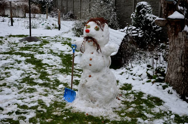 雪人带着一个疯狂而勇敢的粉丝的鬼脸 手拿着照明弹 家庭花园里的小雪和长满了皱纹的草坪 孩子们用铲子和耙来收集材料 — 图库照片