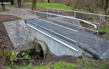 Parkta yeni yenilenmiş bir köprü. Küçük bir derenin üzerinde. Akış hatlarından geçen boruları olan beton yapılar. Asfalt yüzey, metal levha çinko damlatma tepsisi. Boru korkulukları, çimen, bahçe, bisiklet