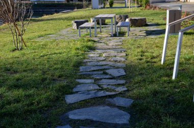 Stone slabs in the lawn, floor in the lawn shape of the rectangle camp site with a table and a urban bench, a lake. large flat blocks, picnic, fireplace, barbecue, park, boulder. sunny day