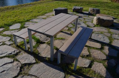 Stone slabs in the lawn, floor in the lawn shape of the rectangle camp site with a table and a urban bench, a lake. large flat blocks, picnic, fireplace, barbecue, park, boulder. sunny day