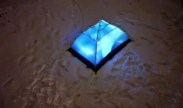 a man in outdoor clothes will sleep in a blue tent directly on snow. support equipment for nature, experience an adventure in wilderness not far from cottage. lights up with a flashlight inside.