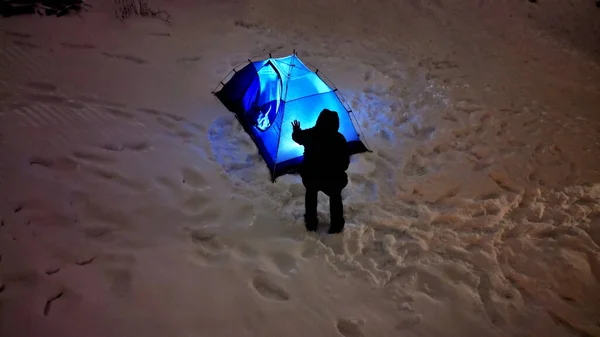 a man in outdoor clothes will sleep in a blue tent directly on snow. support equipment for nature, experience an adventure in wilderness not far from cottage. lights up with a flashlight inside.