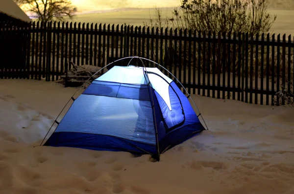 tourist in outdoor clothes will sleep in a blue tent directly on snow. support equipment for nature, experience an adventure in wilderness not far from cottage. lights up with a flashlight inside.