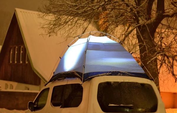 tourist in outdoor clothes will sleep in a blue tent directly on snow. support equipment for nature, experience an adventure in wilderness not far from cottage. lights up with a flashlight inside.