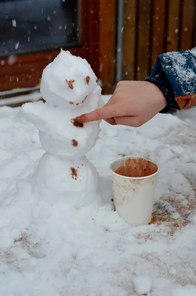 the boy draws the snowman's eyes and mouth with a finger dipped in hot chocolate, he also has buttons. fun painting on snow with brown paint with your hands, paper cup of coffe, cafeteria, table