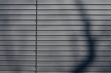 gray building is lined with square panels. metal sheet wall cladding with scalloped design. corrugated sheet, dark gray, riveted. horizontal stripes, shadows, corner, fence, aluminium, sheet