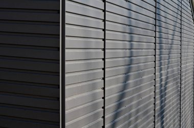 gray building is lined with square panels. metal sheet wall cladding with scalloped design. corrugated sheet, dark gray, riveted. horizontal stripes, shadows, corner, fence, aluminium, sheet
