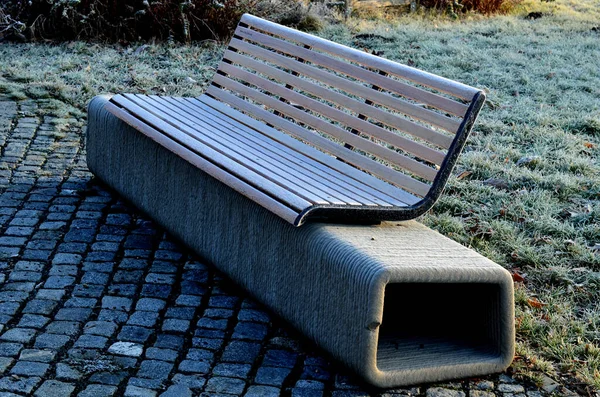 park bench has a concrete tube base flattened on the sides. the bench is printed on a 3d printer from concrete. the cement mass is extruded into a spiral. there is a wooden seat and backrest on top
