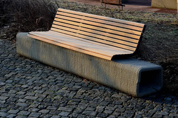 park bench has a concrete tube base flattened on the sides. the bench is printed on a 3d printer from concrete. the cement mass is extruded into a spiral. there is a wooden seat and backrest on top