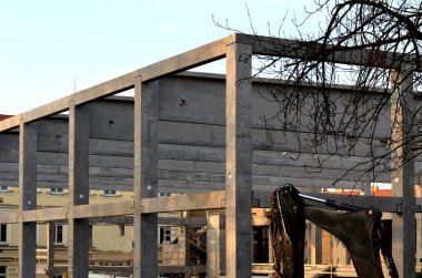 Endüstriyel depo binasının beton yapısı. Karıştırıcının beton besleyici kolu kirişlerin ve kolların üzerine eğiliyor. Vinçte boru taşıyıcısı. Kazıcı, traktör, sarı.
