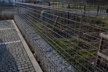Gabion teli sepeti, kafesler. Doğal endüstriyel görünümüyle ön bahçede çit var. Galvanize kablodan yapılmış. Katlanmış taşlarla dolu istinat duvarı. Gürültü bariyeri yerleşim alanını koruyor.