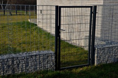 Gabion teli sepeti, kafesler. Doğal endüstriyel görünümüyle ön bahçede çit var. Galvanize kablodan yapılmış. Katlanmış taşlarla dolu istinat duvarı. Gürültü bariyeri yerleşim alanını koruyor.