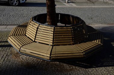 Ağacın gövdesini pas geçen daha küçük tahtalardan oluşan yuvarlak banklarla çevrili, zemin karenin üzerine dikilmiş ağaç kaseleri. Çiçek saksısının asfaltları kademeli olarak granit küplerdir.