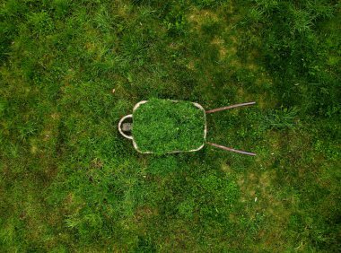 Hasır şapkalı bahçıvan çim biçme makinesinin sepetini el arabasına attı. Güzel yeşil bahçe bakımı. Bir adam meyve bahçesinin yüzeyine bakan bir araba sürüyor.