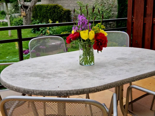 stock image terrace with a vase of meadow flowers on a table in a garden or restaurant. peonies and irises together with yellow tulips in a cylinder-shaped glass container. fresh colors of flowers plucked from