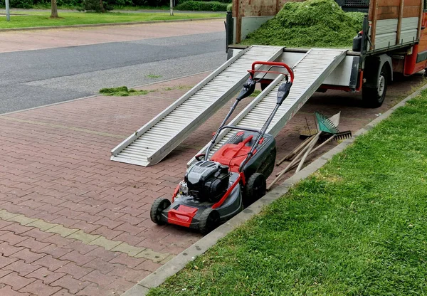 a truck loaded with cut grass. ramps for large mowers made of aluminum profiles. a lawnmower is waiting ready to work in the parking lot of a shopping center. red