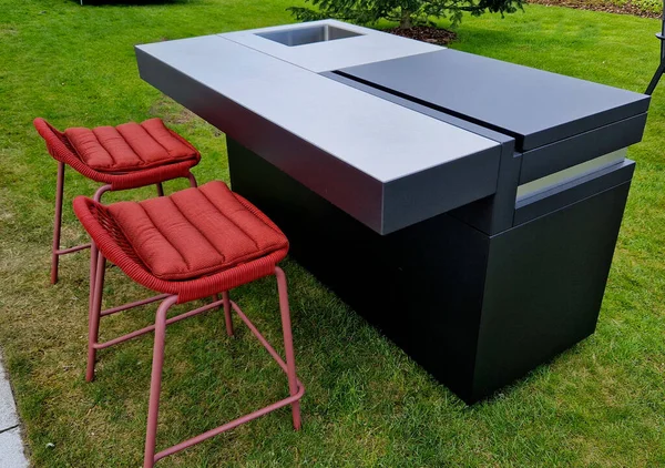 stock image outdoor kitchen with bar stools. metal gray cabinet with gas grill and stainless steel sink. the box is a portable design gray matte sheet. the upholstery of the two chairs is red, textile.