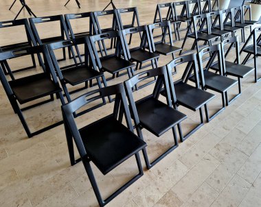 Koridorda plastik katlanır sandalyeler. Ders dinleyicileri ya da etkinlik sunucuları. Yan yana sıralanmış sıralar. İç kısımlarında bej-kahverengi asfalt var.