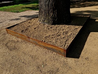 Parkın ortasındaki yaşlı ağacın korunması için. Kare şeklindeki paslanmaz çelik kaldırım kaldırımın çakıl ve kum yüzeyinin biraz üstündedir. Arazi yukarıda yükseldi.