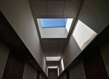 Başının üstünde dikdörtgen bir pencere var. Koridorun tamamını doğal ışıkla aydınlatıyor. Pencere dikdörtgen bir tavan penceresi. Duvarlar bembeyaz.