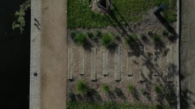 Park ziyaretçileri için iskele. İki adım ona götürür. Suyun üzerinde kalaslar var. Tekneler için rıhtımı olan bir gölet. Bir dikdörtgenin kat planı. Terasta parmaklık yok. Taş, yeşil, yosun, tekne, çimen.