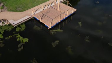 Park ziyaretçileri için iskele. İki adım ona götürür. Suyun üzerinde kalaslar var. Tekneler için rıhtımı olan bir gölet. Bir dikdörtgenin kat planı. Terasta parmaklık yok. Taş, yeşil, yosun, tekne, çimen.