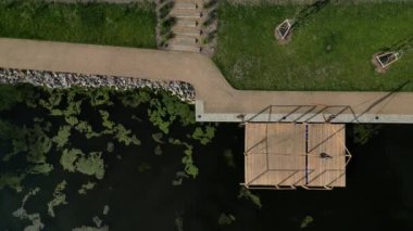 Park ziyaretçileri için iskele. İki adım ona götürür. Suyun üzerinde kalaslar var. Tekneler için rıhtımı olan bir gölet. Bir dikdörtgenin kat planı. Terasta parmaklık yok. Taş, yeşil, yosun, tekne, çimen.