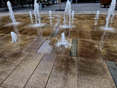 Kaldırımda kaldırıma yığılmış kare granit kaldırım taşları, sırayla püskürtülmüş su püskürtüleri. Su özellikleri doğrudan park sırasındaki kaldırımlı meydandan ortaya çıkıyor. Islak kiremitler tahta kaldırım, bebeğim, kep
