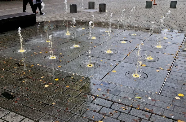 square granite paving stacked in a strip in sidewalk,boy, water sprays with alternating spray heights . water features emerge directly from cobbled square in park row. wet tiles, led, yellow light
