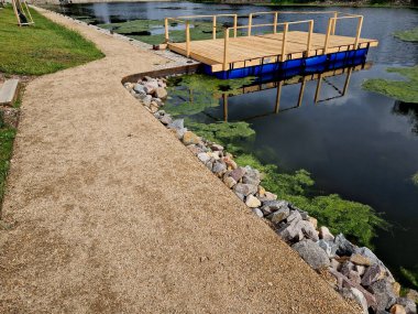 Park ziyaretçileri için iskele. İki adım ona götürür. Suyun üzerinde kalaslar var. Tekneler için rıhtımı olan bir gölet. Bir dikdörtgenin kat planı. Terasta korkuluk yok, taş, yeşil, yosun, tekne, ördek otu, çimen