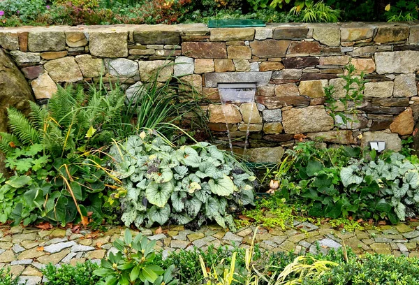 trickle of water flows from a joint in stone wall along a stainless steel sheet into a pond with perennials and ferns around it. beautiful folded walls in park and garden will be made by our company