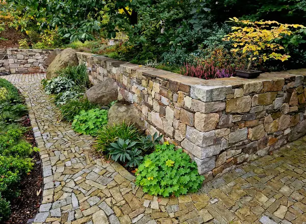 trickle of water flows from a joint in stone wall along a stainless steel sheet into a pond with perennials and ferns around it. beautiful folded walls in park and garden will be made by our company