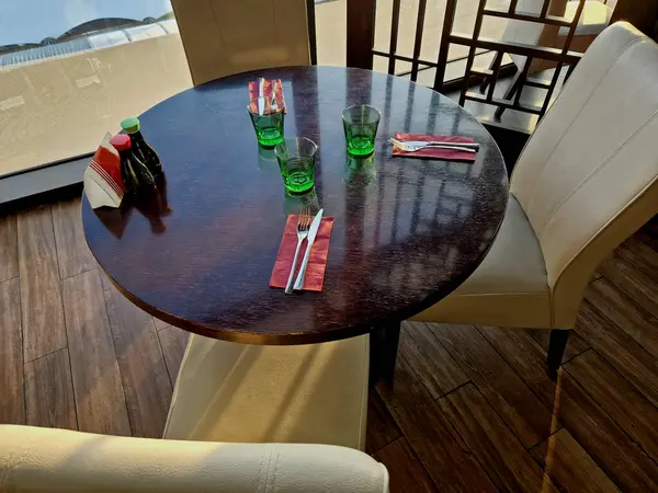 lacquered circle-shaped table with leather kitchen chairs and bistro cutlery is prepared on the table top. red paper napkins and green drinking glasses. bright modern interior