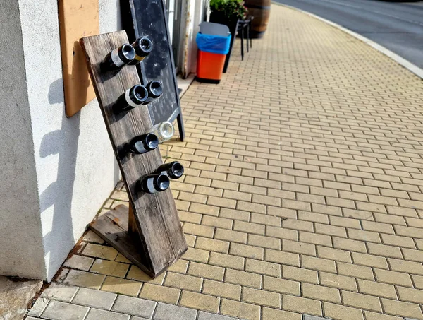 the wine shop on the street is equipped with a wine rack made of a piece of board with holes in it and bottles in them face down. they are empty and serve as an advertising banner on the sidewalk