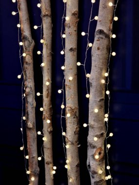 Koyu mavi arka plandaki ışık zincirleri gece yıldızlı bir gökyüzünü andırıyor. Noel için aydınlatılmış süslemelerle sergi standı, dal, daire, huş ağacı kabuğu, beyaz, 