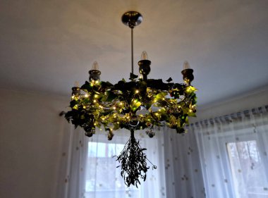 Sekiz kollu eski bir avize. Gümüş renkli, ladin konisi şeklinde ampulleri ve altta bir topu var. Perdeli beyaz oda. Alt manzara. Mum şekilli, örümcekli, sarılan, öne çıkan, krom