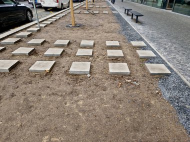 Basamaklar çiçek tarhının ortasına çıkar. Ağacın etrafında damlayan bir boru var. Toprak yüzeyinde otomatik sulama hortumu, dama tahtası ızgarasında kare beton fayanslar