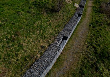 Gabion boruları olan duvarları yolun kenarındaki hendeğe boşaltma suyu olarak kullanıyor. Tahta banklar yerleştirilmiş, parktaki bir duvara monte edilmiş.