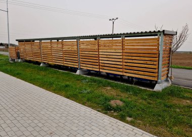 Otobüs durağı olan pergola çatısı. Çöp kutusuyla birleştirilmiş. Plastik, kağıt, cam renkli kutulara ayrılmış. Tahta levhalar, mavi gökyüzü, bulutlar, süpürge, mala, kaldırım, sokak