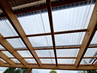 Ahşap pergola ve pleksiglass çatılı teras. Sarmaşıklar geriliyor, kirişlerin altında sürünüyorlar. Bahçe ya da park. Alkolsüz duvar şarabı bölgesinde oturuyor. Kırsal restoran Fransa, kiriş, küre, lamba