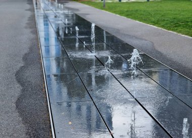 Kaldırımdaki bir şeride yığılmış kare granit kaldırım, sırayla püskürtülmüş su püskürtüleri. - Evet. Su özellikleri doğrudan park sırasındaki kaldırımlı meydandan ortaya çıkıyor. ıslak kiremitler tahta kaldırım