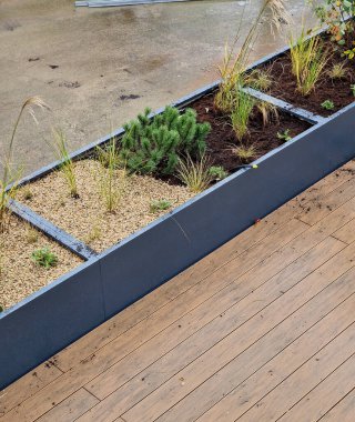 Tahta bir terasta gri plastikten yapılmış kendi kendine sulandırıcı kaplar otlar ve cüce formunda çam ağaçları ekilmiş. Duvarda bir sıra dikdörtgen çiçek saksısı.