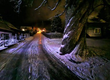 christmas atmosphere snowy road and old trees. however, someone decorated a small spruce with ice chains. a snowdrift by a lamp on the street clipart