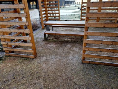 Duvarları ve çatısı çizgili ahşap pergola. Tırmanma bitkileri için çardak görevi görüyor. Kumlu bej yüzey. Tel banklarla parktaki patikalar. Vidaları ve iplikleri olan temel çubuklar, kış