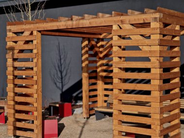 Duvarları ve çatısı çizgili ahşap pergola. Tırmanma bitkileri için çardak görevi görüyor. Kumlu bej yüzey. Tel banklarla parktaki patikalar. Vidaları ve iplikleri olan temel çubuklar, kış