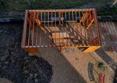 Duvarları ve çatısı çizgili ahşap pergola. Tırmanma bitkileri için çardak görevi görüyor. Kumlu bej yüzey. Tel banklarla parktaki patikalar. Vidaları ve iplikleri olan temel çubuklar, kış